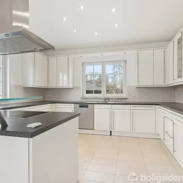 Et moderne køkken med hvide skabe og sorte bordplader, centralt placeret kogeø med emhætte, indbyggede ovne i højre side, flisegulv og stort vindue med udsigt til have.