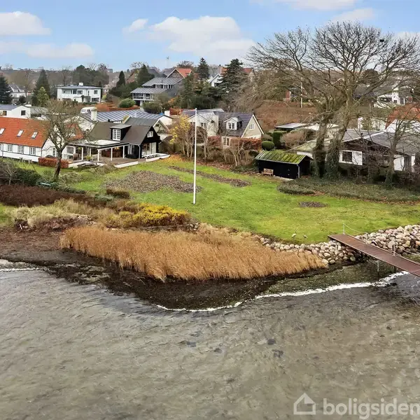Huse ved kystlinje med en træbro strækker sig over vandet; omgivet af grønne områder, træer og andre boliger i baggrunden.