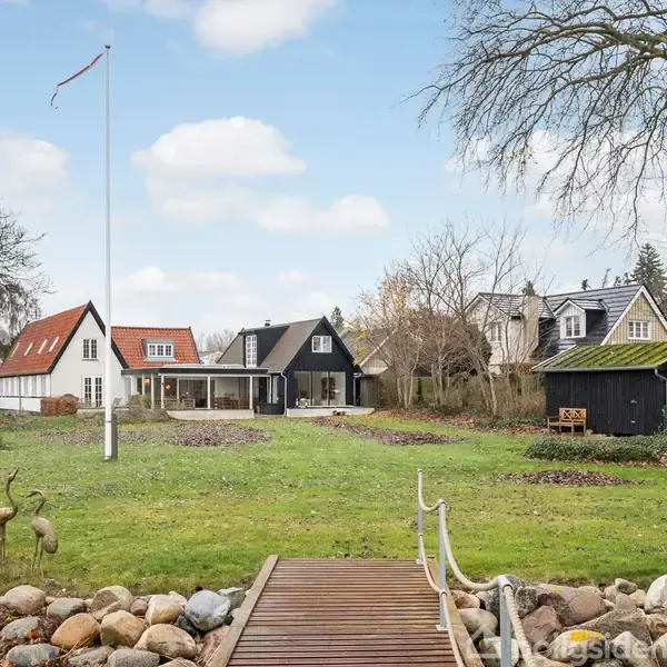 Græsplæne strækker sig foran flere huse i klassisk stil. Huse i baggrunden, omgivet af træer og buske. Træbro krydser en stensætning, hvor kranefigurer pynter haven.