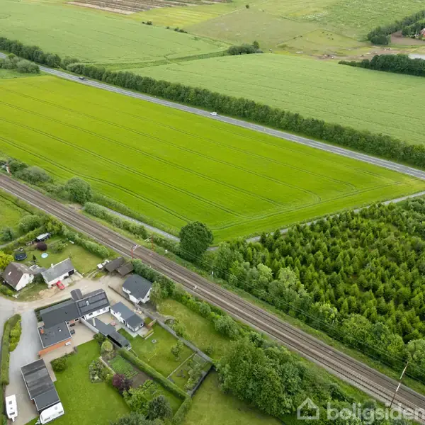 Landsbyhus ligger stille i et landligt område, omgivet af en stor grøn mark og en jernbanespor. Spredte huse og træer omkranser markerne, skaber en fredelig landlig scene.
