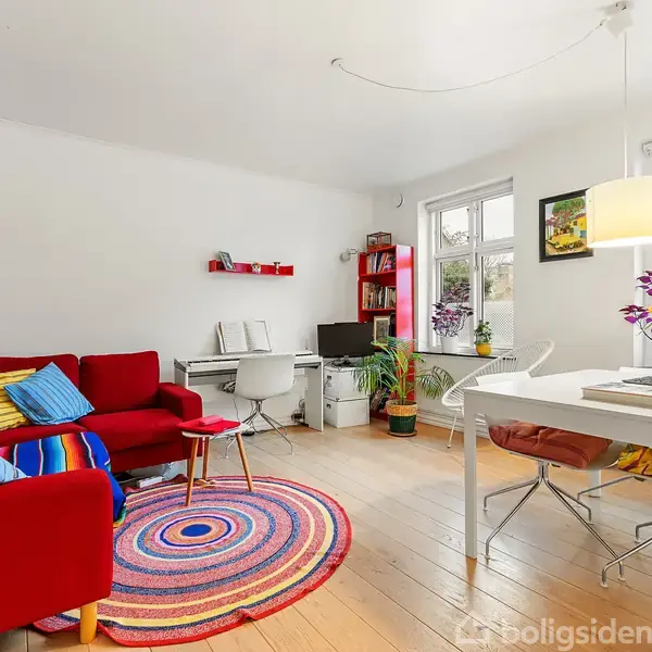 Rød sofa med farverige puder står i en stue. Ved siden af er et skrivebord med computere. Et rundt tæppe og et bord med blomster understreger det lyse rum med store vinduer.