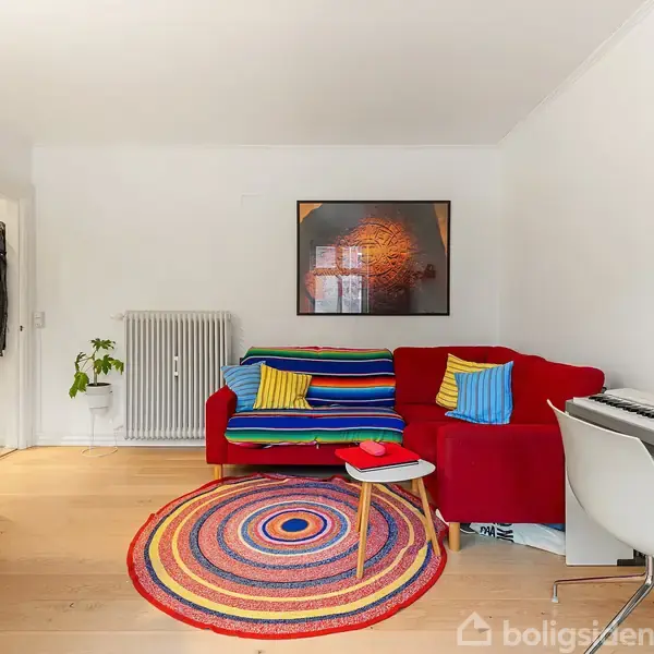 Rød sofa med farverige puder står mod en hvid væg ved siden af et klaver. Rundt tæppe på trægulvet. En dør med hængende jakke og en potteplante ses i baggrunden.