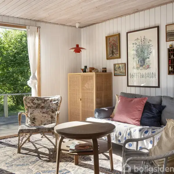 Stue med grå sofa, puder og to stole foran et rundt sofabord. Vægge med billeder og en hylde. Glasdør åbner til en træterrasse omgivet af grønne træer.