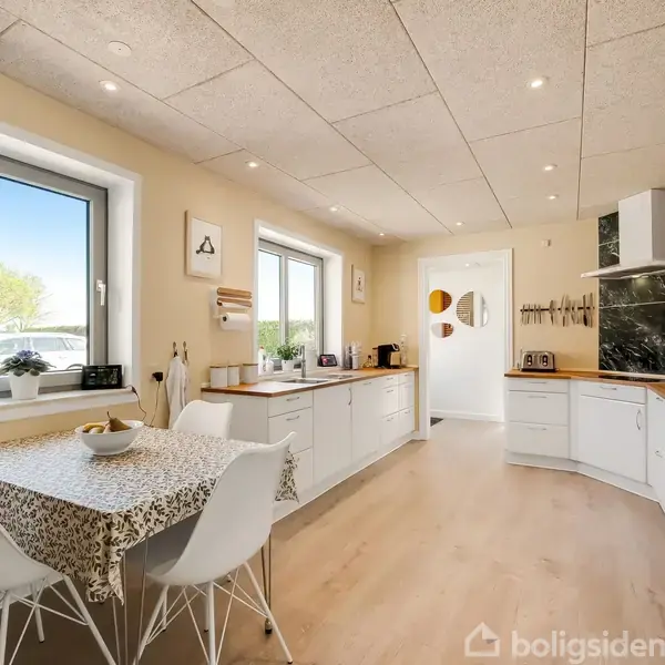 Køkken med hvidt interiør. Bord med fire stole står ved vindue. Køkkenbordet har træoverflade og moderne apparater. Lyse vægge og lofter med indbyggede spots. Udsigt til natur udenfor.