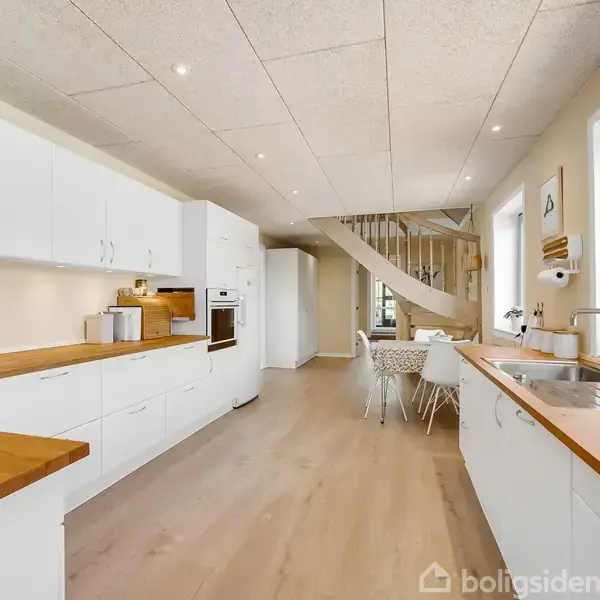 Et lyst køkken med hvide skabe og træbordplader står stille. Det åbner op mod en spiseplads og en vindeltrappe. Et vindue til højre bringer naturligt lys ind.