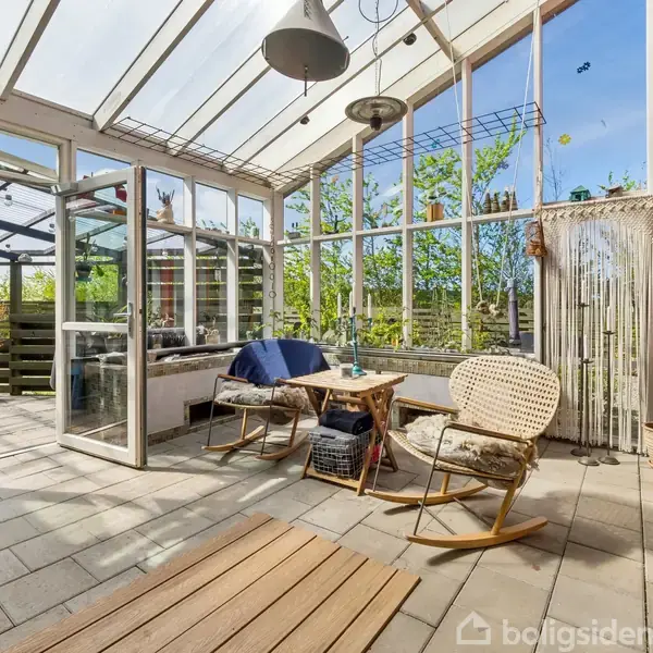 Glasoverdækket terrasse med to stole omkring et bord, omgivet af planter. Åben glasdør fører ud til en have. Lys hænger fra loftet, skaber en behagelig atmosfære.