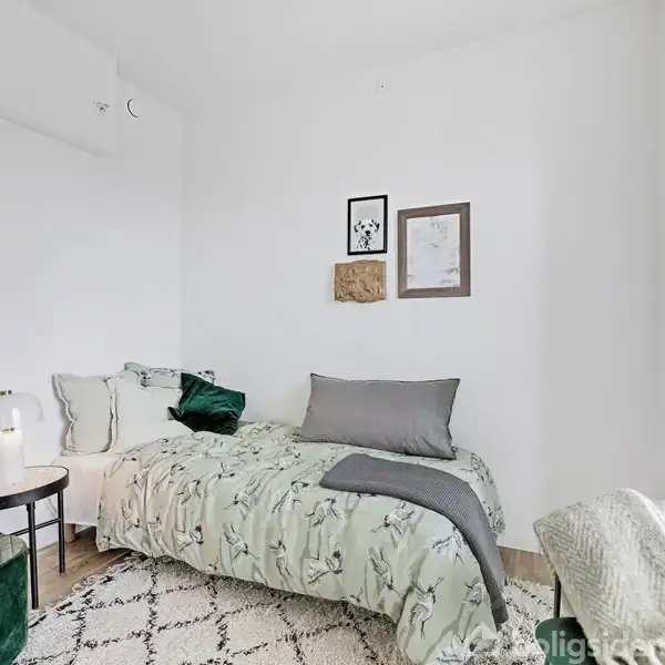 En enkeltmandsseng med blomstret sengetøj står i et hjørne, omgivet af hvide vægge og dekoreret med vægkunst. Ved siden af sengen står et lille rundt bord med en lampe.