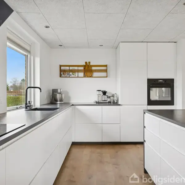 Et moderne køkken med hvide skabe, sorte bordplader og en indbygget ovn. Et vindue til venstre giver naturligt lys, og køkkenredskaber står på bordet.