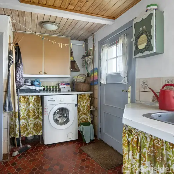 Vaskemaskine står stille i et lille, retro-stil vaskerum med farverige gardiner og fliser. Et gråt skab hænger på væggen. Håndvask og køkkenartikler til højre.