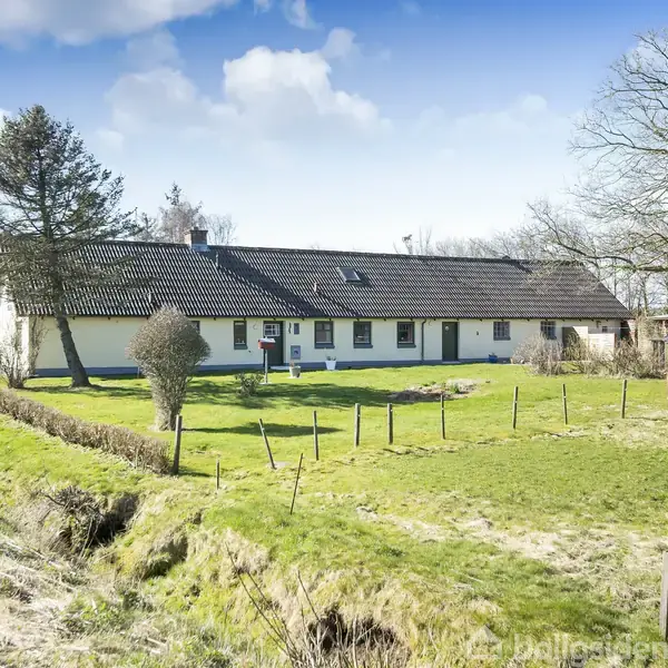 Et langstrakt hus med mørkt tag og hvide vægge står i en grøn mark, omgivet af træer og en lav hegn. Himlen er blå med spredte skyer.