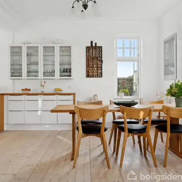 Et moderne køkken med spisebord omgivet af fire stole. Der er lyse trægulve, hvide skabe med glasdøre og grønne planter ved vinduet, der giver udsigt til en have.