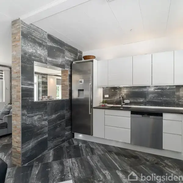 Køkken med hvide skabe og rustfri stål-apparater, herunder køleskab og opvaskemaskine. Rummet har marmor-lignende vægge og gulve. I baggrunden er en stue med en grå sofa og store vinduer.