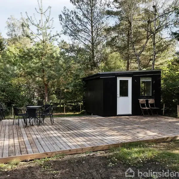 En træterrasse med sorte havemøbler foran et sort skur i en skovklædt have. Skuret har en dør og et sidebord med to stole.