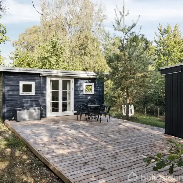 Et sort træhus står på en træterrasse med et rundt bord og fire stole. Omgivet af frodig skov giver huset en fredelig atmosfære.