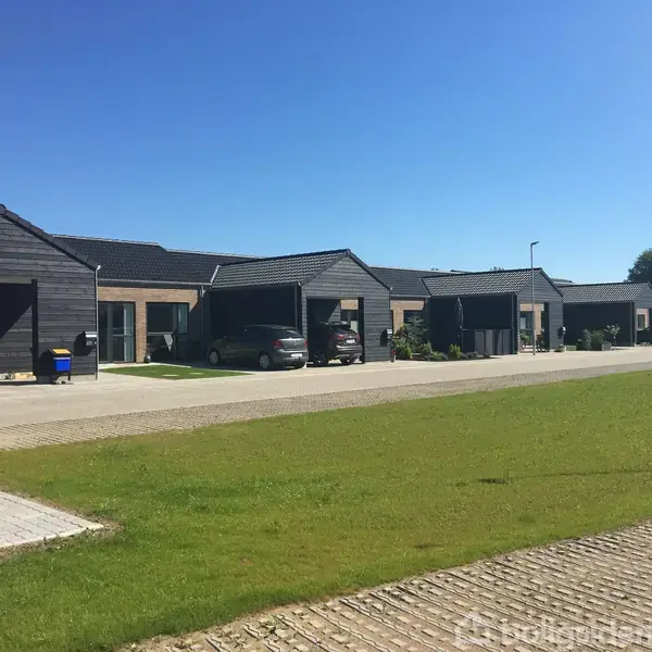 Rækkehuse med sorte tage står i en række. Biler er parkeret i carporte foran husene. Omgivet af en grøn græsplæne og en klar blå himmel.