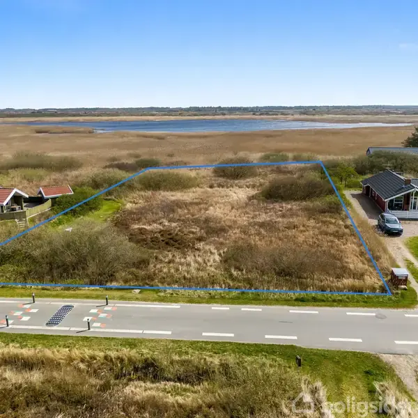 Byggetomt med højt græs, afgrænset af blå linjer, ved siden af huse og en vej i forgrunden. I baggrunden ses en sø og fladt naturlandskab.