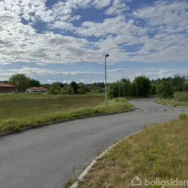 En asfalteret vej drejer blødt til højre, omkranset af græs og buske, med en enkelt lygtepæl og boliger i baggrunden under en delvist overskyet himmel.