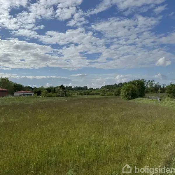 En stor, åben mark strækker sig mod horisonten under en delvis skyet blå himmel. I baggrunden ses nogle huse og træer, mens en ensom gadelygte står til højre.