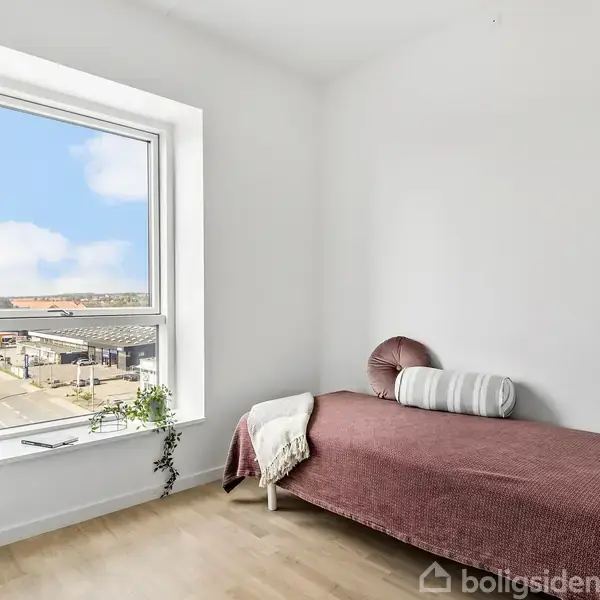 En enkeltseng er placeret ved en hvid væg med puder og brun sengetæppe. Den står nær et vindue med udsigt over en by og blå himmel.