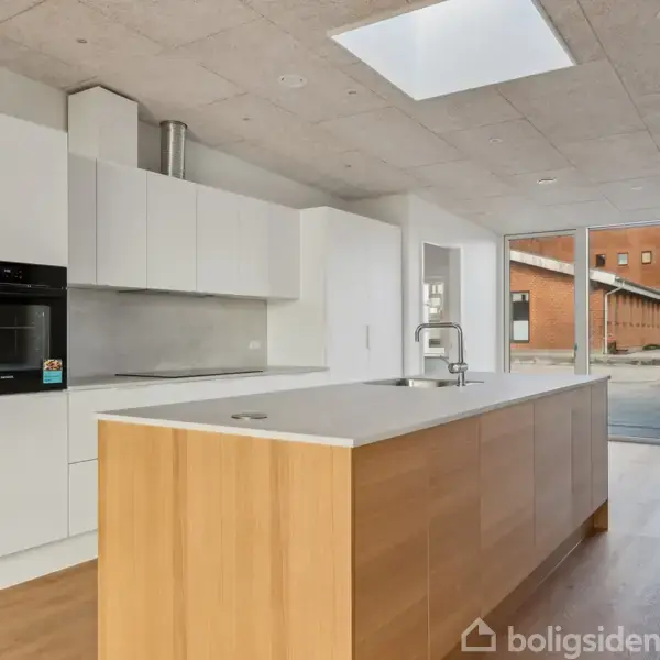 Køkkenø med træpaneler og indbygget vask står i et moderne, lyst køkken med hvide skabe og ovn. Store vinduer viser en udsigt til en gård med bygninger.