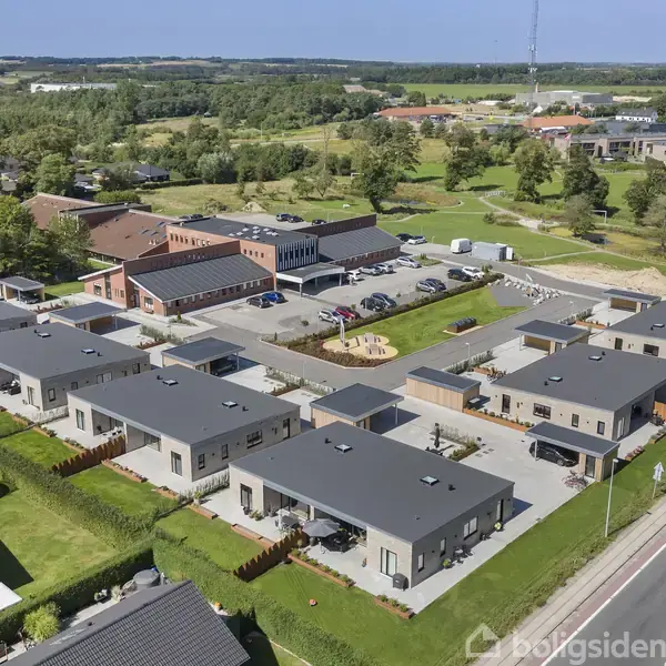 Flade tage på rækkehuse set fra luften, omgivet af grønne haver og en vej til højre. I baggrunden ses en parkeringsplads og flere større bygninger samt landlige omgivelser.
