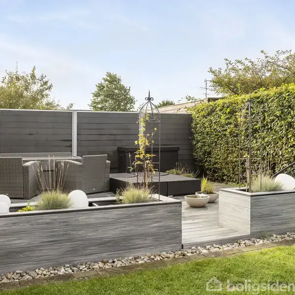 Terrasseområde med moderne havemøbler arrangeret på et trædæk. Omkring er der grå hegn og en tæt hæk, med græsplæne og beplantede blomsterbede i forgrunden.