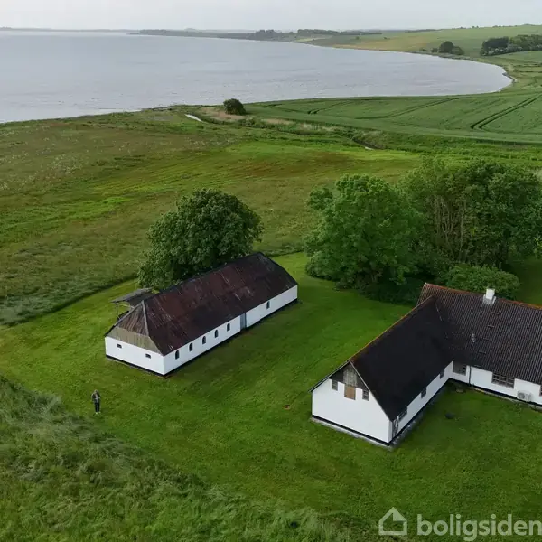 To hvidkalkede bygninger med brune tage står på en grøn græsplæne. De er omgivet af træer med en sø i baggrunden og marker i det fjerne.