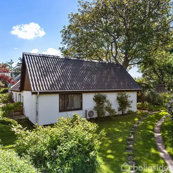 En hvid bygning med skråt tag står i en frodig have, omgivet af buske og træer. En stenbelagt sti snor sig gennem græsplænen i solrigt vejr.