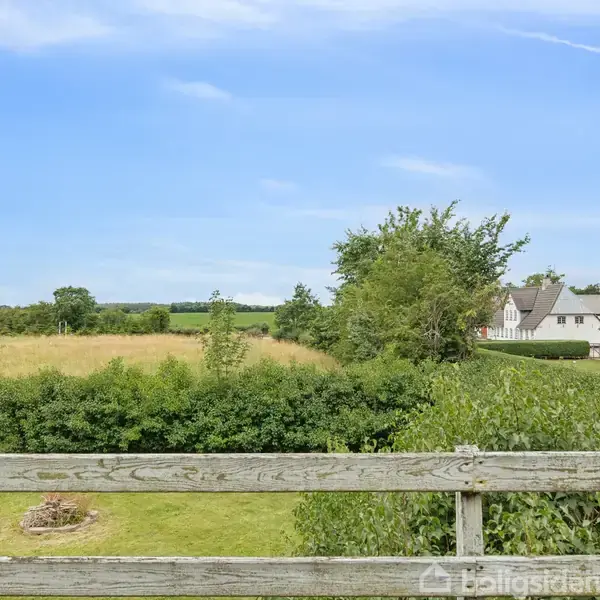 Landskab ses fra en lys træterrasse. En åben mark og grønne buske dominerer forgrunden, mens et par huse står i baggrunden under en klar, blå himmel.