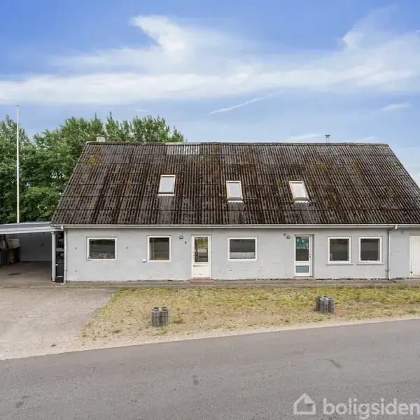 En gråmalet bygning med skråtag og flere ovenlysvinduer står ved en asfaltvej. Til venstre en carport med brædder. Træer i baggrunden fuldender landskabet.