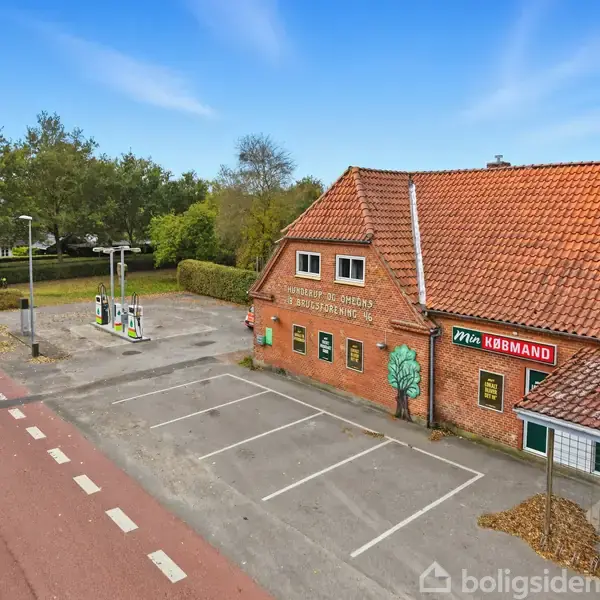 Rødt murstensbygning med skiltet "Min Købmand" og "Nyborgvej 950 Ørbæklunde, FDB's Mindelund" ved siden af en vej og en benzinstation, omgivet af grønne træer.