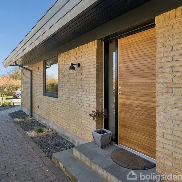 Huset har en gul murstensfacade med en trædør og to vinduer. Der er en flisebelagt gangsti omgivet af grus og grønne buske.