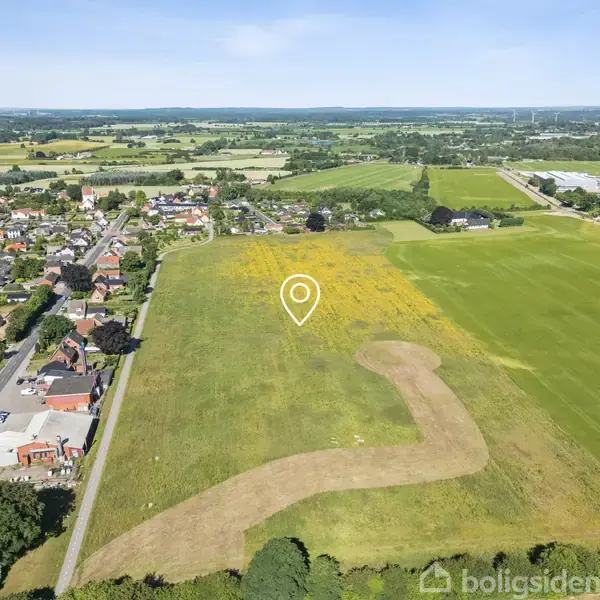 Grønt græsareal med en markør over et felt; beliggende ved en landevej. Naboer udgør et lille boligområde på venstre side. Store marker strækker sig til højre og baggrunden.