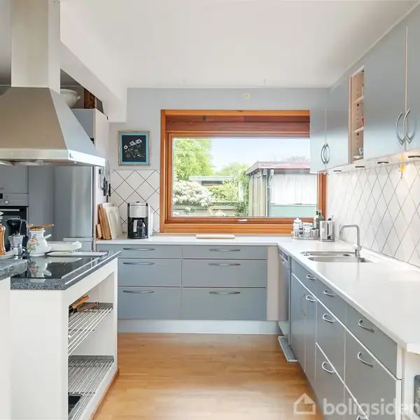 Et køkken med grå skabe og hvid bordplade. Køkkenet har en stor udsugningshætte, et stort vindue med udsigt til en have og hvide fliser med diamantmønster som backsplash.