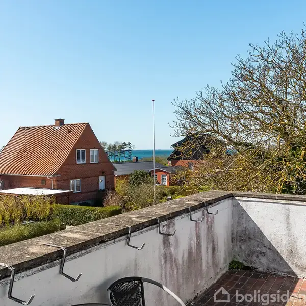 En terrasse med stole og bord. En stor trækrone skygger. I baggrunden ses et gammelt murstenshus og udsigt mod havet under en klar blå himmel.