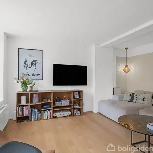 Stue med en moderne sofa i hjørnet, vægmonteret TV over en reol med bøger og planter, samt et rundt sofabord foran. Store vinduer slipper lys ind.