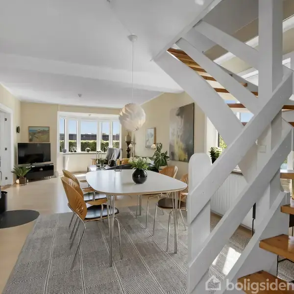 En moderne spisestue med rundt spisebord og stole, omgivet af lysindfald fra vinduer. En trappe fører opad. Rummet er åbent og lyst, med en brændeovn i hjørnet.