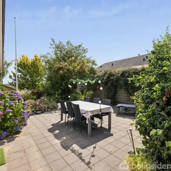 Terrassemøbler står på flisebelagt terrasse med et bord og seks stole. Omgivet af frodige buske og blomster. Huset ses til venstre, og himlen er skyfri.
