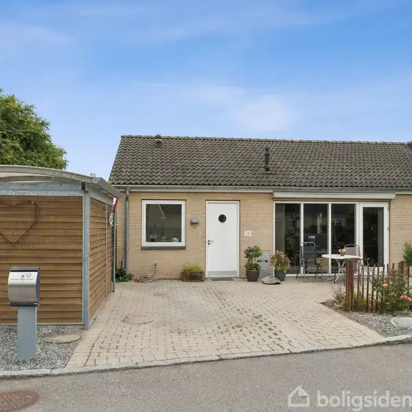 Et enkelt enfamilieshus står i et boligområde, omgivet af en lille have og et hegn. Der er en carport til venstre, og himlen er klar blå.
