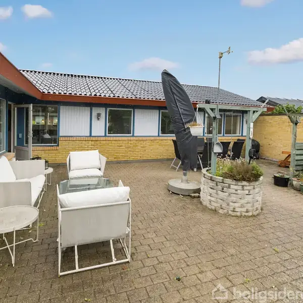 Terrassemøbler med hvide hynder står på en solrig, flisebelagt gårdhave omkranset af en gulstensbygning med store vinduer. Et bord og en plante i midten, omgiven af diverse krukker.