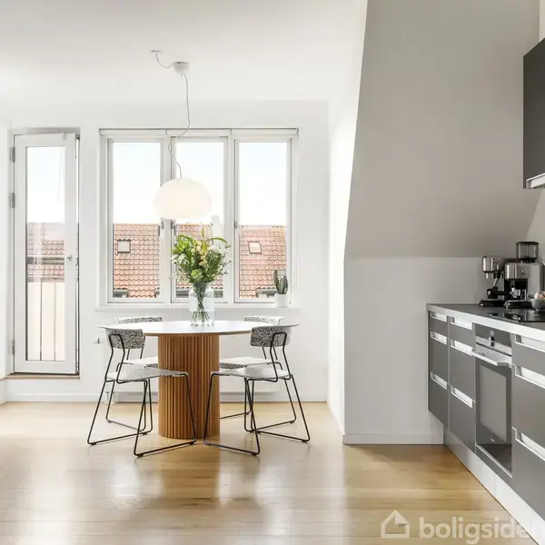 Et moderne køkken har et rundt bord og fire stole, med en vase med blomster ovenpå. Store vinduer giver udsigt til røde tegltage. En køkkenbordsplade løber langs højre side.