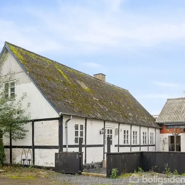 Et hvidt bindingsværkshus med sort træværk står på en gårdsplads. Husets tag er dækket med mos, og der er en grusbelagt forplads omgivet af grønne områder.