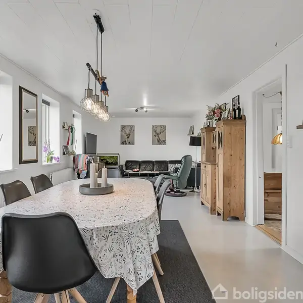 Spisebord med lys dug og sorte stole står centralt i rummet, mens lamper hænger over bordet. Stuen i baggrunden har sofaer og reoler, omgivet af hvide vægge og vinduer.