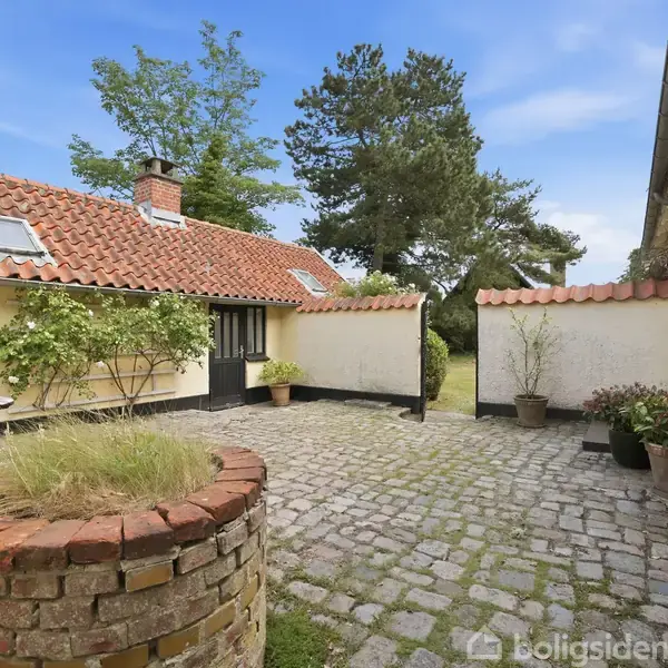 Gårdhave omgivet af murede bygninger med røde teglstenstage; haveborde og planter pryder stenbelægningen. Skyfri himmel og træer i baggrunden.