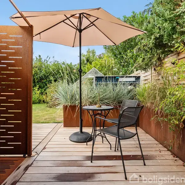 En have med træterrasse, hvorpå der står et bord med to stole under en stor parasol. Omkring terrassen er der planter og træer, og i baggrunden ses en græsplæne.