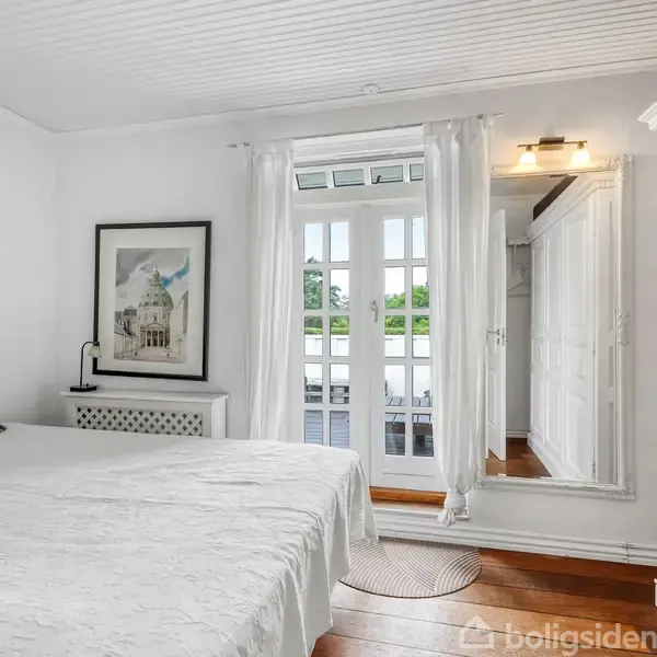 Soveværelse med en stor seng dækket af en hvid dyne; der er puder bagpå. Et billede hænger på væggen. Franske døre fører ud til en balkon med udsigt over grønne træer.