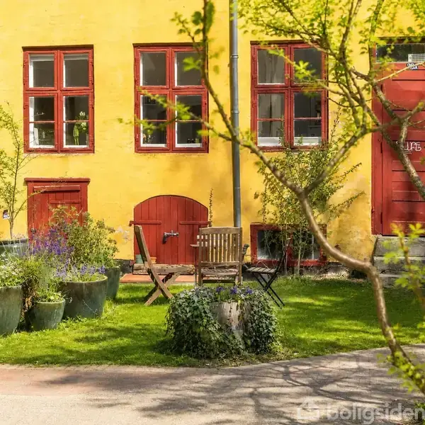 En gul bygning med røde vinduer og døre. En have med krukker, planter og et træ i forgrunden. Træmøbler står på græsset ved siden af bygningen.