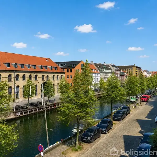 En række biler holder parkeret langs en træbeklædt kanal. En bygning med rødt tag og buede vinduer står til venstre. Lyse blå himmel danner baggrund.