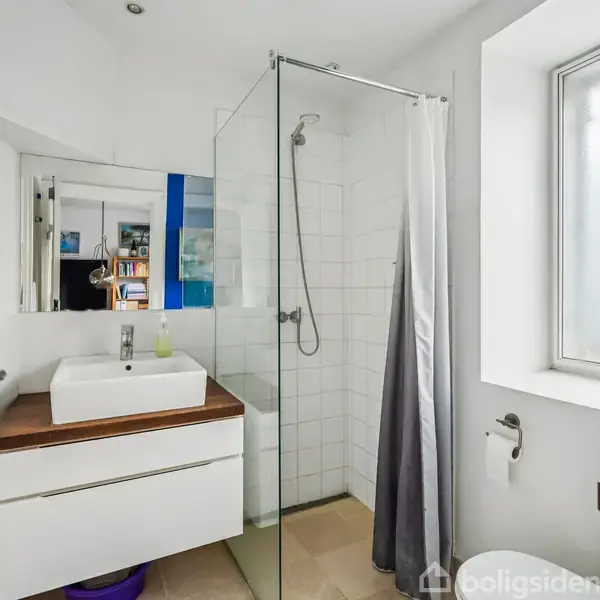 En moderne badeværelsesvask står på et hvidt skab. En glasbruseniche med hvidt flisebelagt væg og en grå badeforhæng ses i nærheden. Et vindue lader lys ind fra højre side.