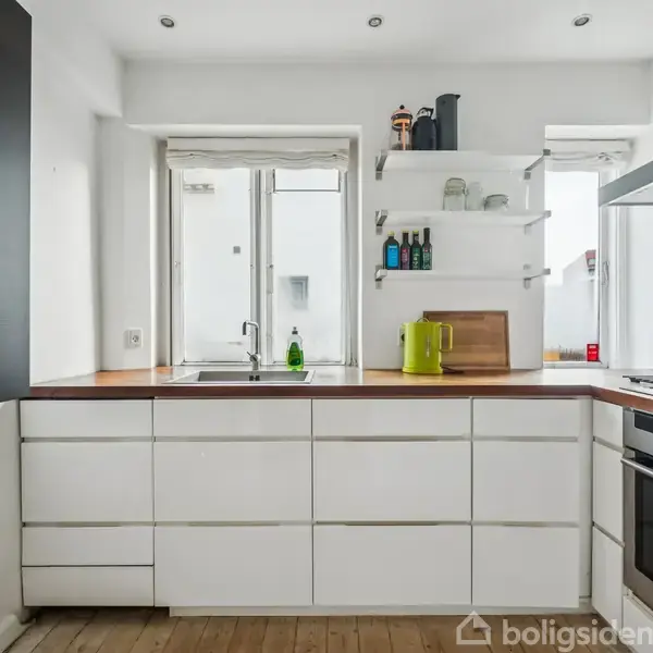 Hvidt køkken med træbordplade; opvaskemiddel ved vask. Gavle, hylder med dåser/flasker, el-kedel. Komfur til højre, trægulv. Vinduer bagved lader lys ind.